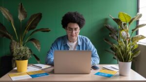 A woman sitting at a desk, looking at a laptop, surrounded by mental health resources and NDIS brochures