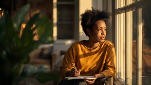 Young woman NDIS participant of African heritage sitting by a sunlit window holding a notebook journal to document functional impact and recovery progress.