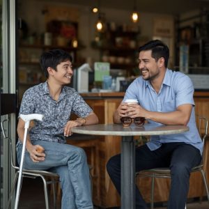 Young adult with vision impairment enjoying coffee at a local café with a SALT Foundation support worker, with a white mobility cane beside the chair, highlighting community inclusion and social connection.