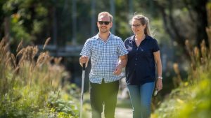 Young adult with vision impairment exploring a community garden using a white mobility cane while walking alongside a SALT Foundation support worker in a community garden setting.
