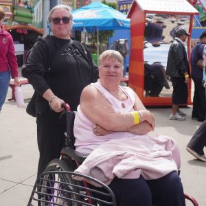 A woman in a pink top sitting in a wheelchair with her arms crossed, smiling alongside another woman in a black shirt standing behind her at the Salt March at Luna Park.