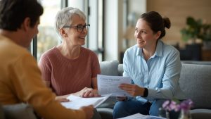 A smiling support worker in a blue shirt discusses a Carers Payment document with an older woman with short grey hair and glasses, while a man in a yellow shirt listens in a comfortable living room setting.