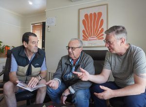 Three older men sitting together indoors engaging in a lively conversation. One man holds a brochure while the other two point and gesture as they talk in a relaxed, friendly setting.